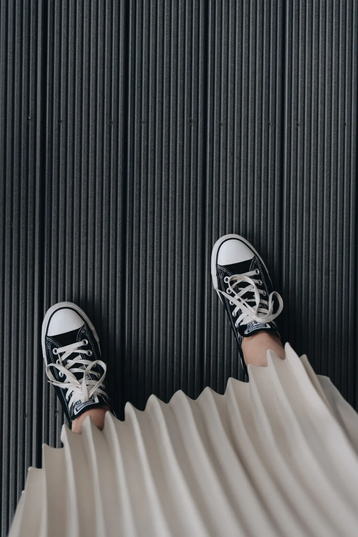 Ujung rok plisket lipit akordeon berwarna krem dan sepasang sneakers kanvas hitam bertali putih, difoto dari atas di lantai bergaris abu tua.