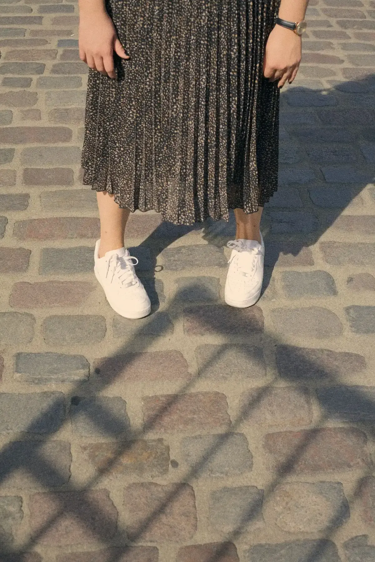 Rok plisket midi bermotif bintik krem di atas dasar cokelat tua dipadukan dengan sneakers putih dan jam tangan, difoto dari pinggang ke bawah di jalan berbatu.