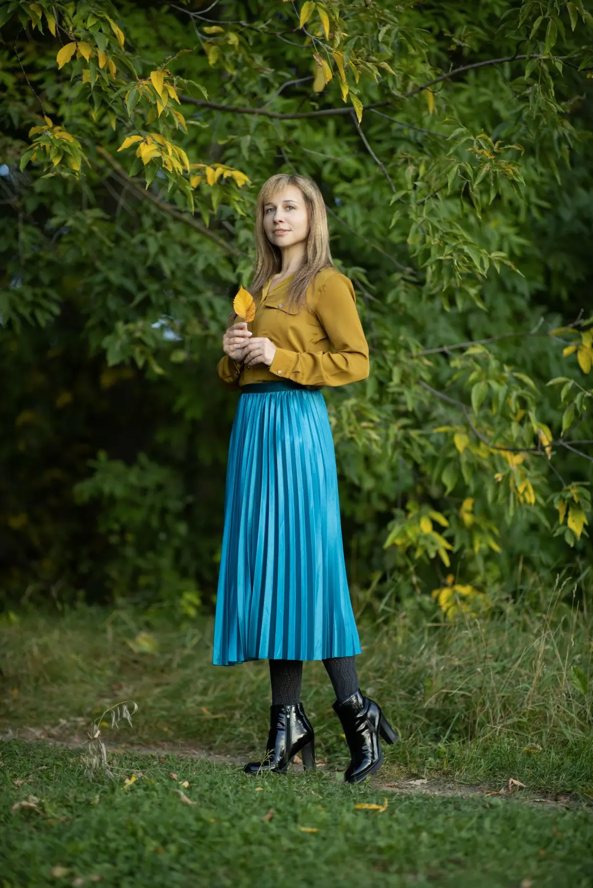 Perempuan memakai blus kuning mustard yang dimasukkan ke rok plisket midi biru toska berkilau, stoking hitam, dan ankle boots hitam, berdiri di taman berdaun hijau dan kuning.