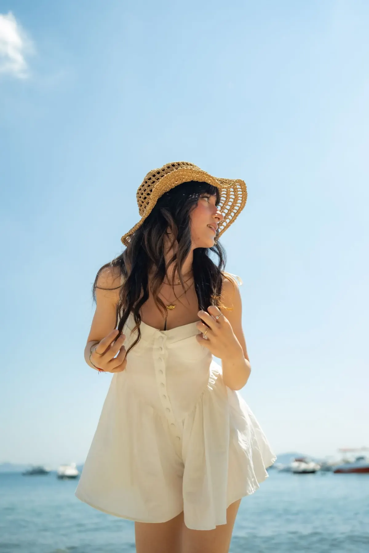 Perempuan berambut panjang memakai romper krem bertali tipis berkancing depan, kalung, dan topi jerami rajut, berdiri di tepi laut dengan langit cerah.