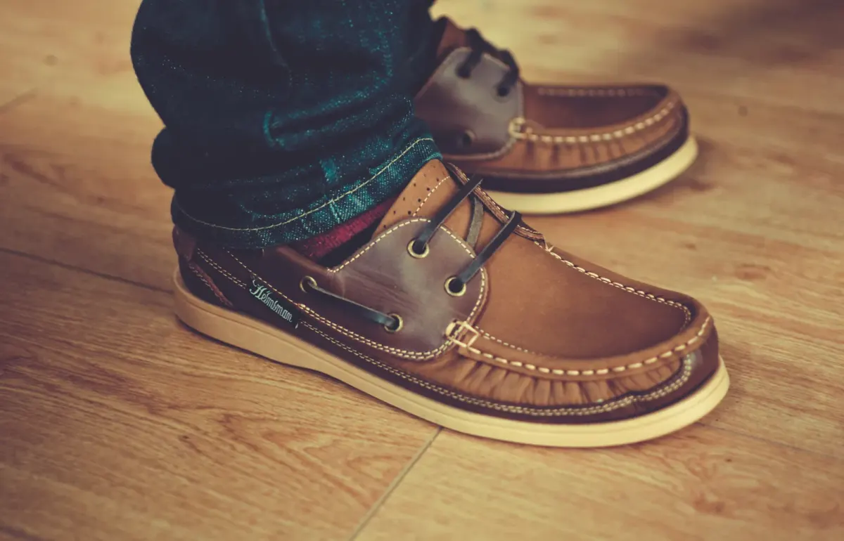 Sepasang sepatu kulit cokelat bertali dengan jahitan terlihat dan sol karet krem, dipakai dengan jeans biru gelap yang ujungnya digulung, di atas lantai kayu.