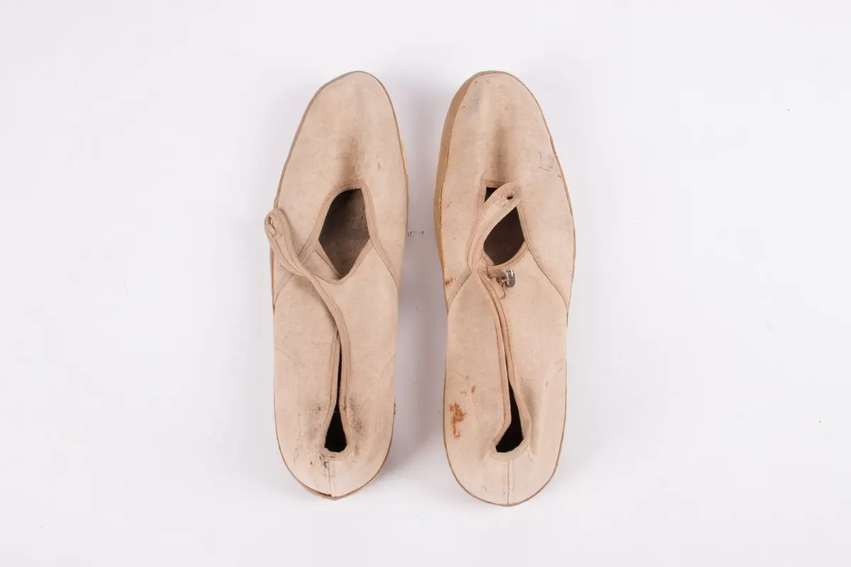 Sepasang sepatu kanvas krem model Mary Jane dengan tali pengait di punggung kaki dan sol karet, difoto dari atas di latar putih sebagai benda koleksi museum.