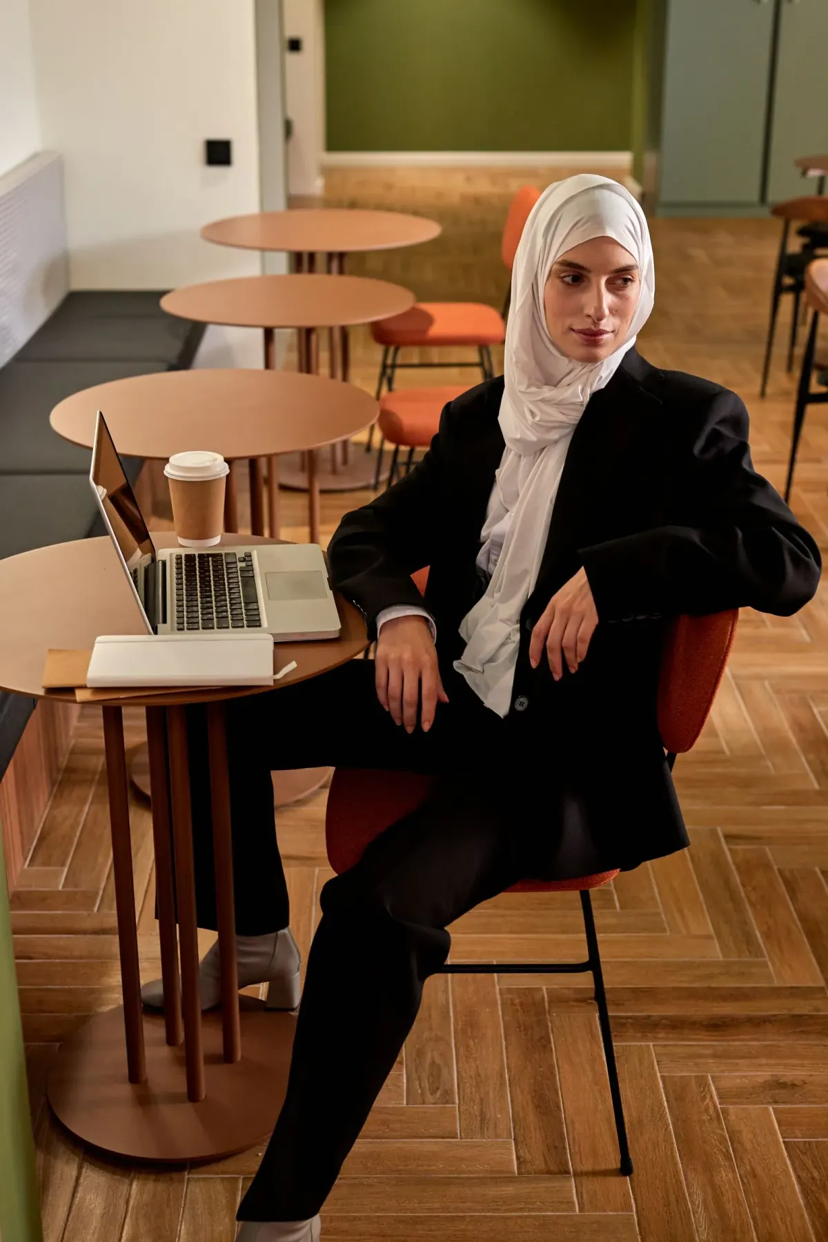 Perempuan berhijab pashmina putih panjang memakai jas dan celana hitam dengan manset kemeja putih terlihat, duduk di kursi kafe dengan laptop dan gelas kopi di meja.
