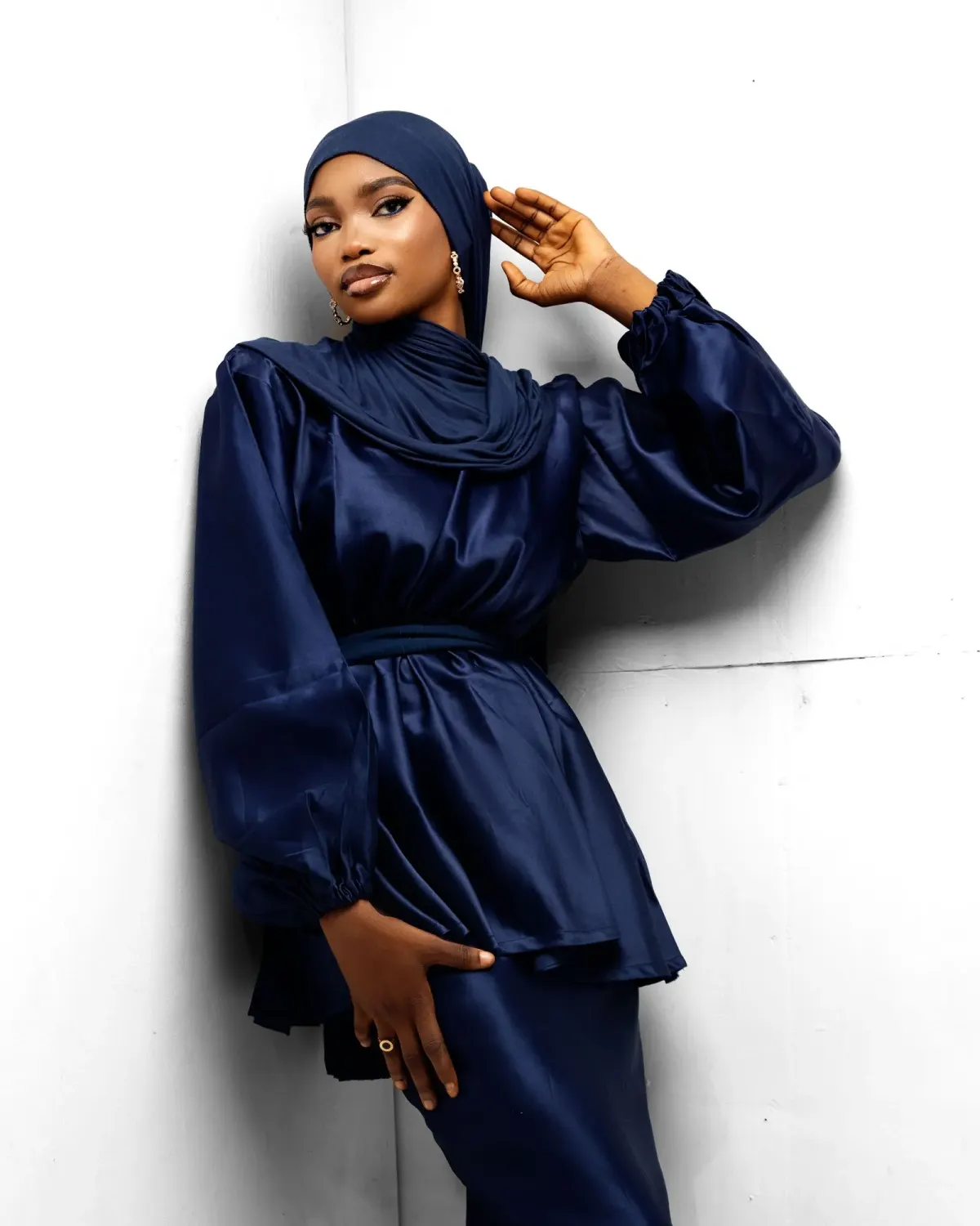 Perempuan memakai atasan satin navy berlengan balon dengan sabuk kain, rok navy, dan jilbab navy yang dililit di leher, bersandar di dinding putih.