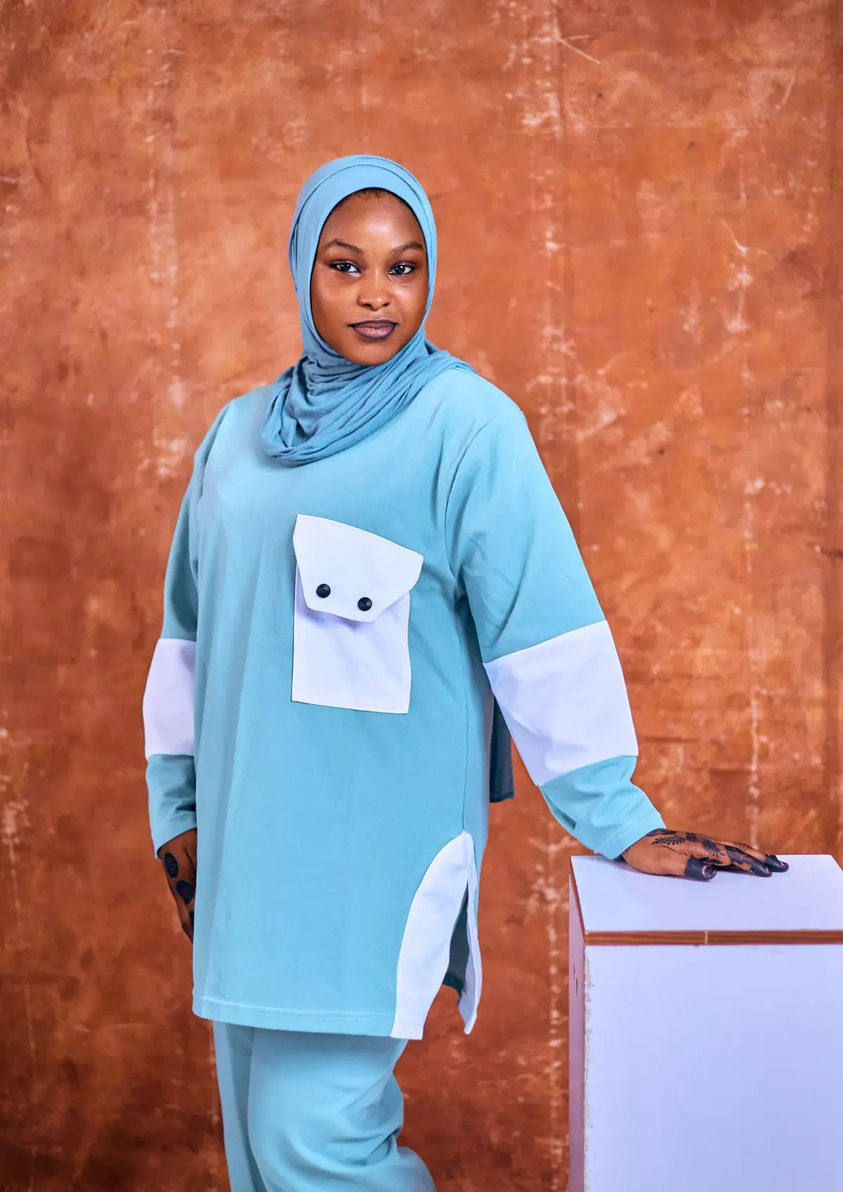 Perempuan berhijab tosca muda memakai atasan longgar lengan panjang tosca dengan panel lengan, saku dada, dan belahan samping berwarna putih, serta celana tosca, berpose di studio berlatar cokelat.