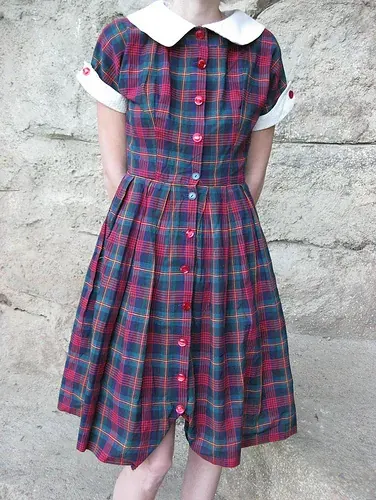 Dress kemeja vintage bermotif kotak merah dan hijau dengan kerah putih bulat, kancing merah dari leher sampai ujung rok, dan rok berlipit, dipakai berdiri di depan dinding batu.