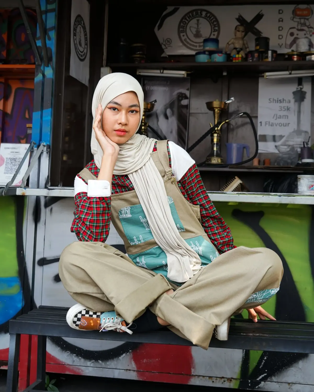 Perempuan berhijab krem duduk bersila di bangku depan kios bergrafiti, memakai kemeja kotak-kotak merah dan atasan putih yang dipakai berlapis, overall khaki bertambal motif tosca, dan sneakers bermotif kotak-kotak.