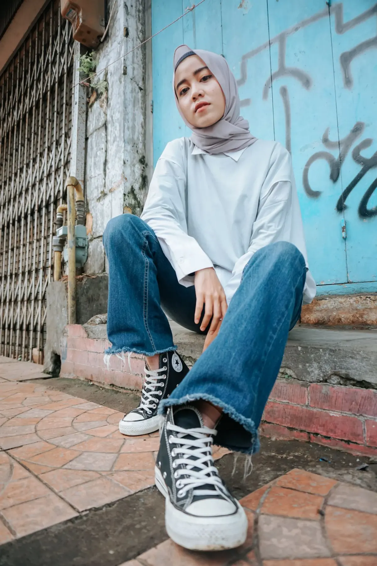 Perempuan berhijab abu-abu duduk di undakan depan pintu biru bercoretan, memakai kemeja putih longgar, jeans biru berpotongan lebar di bawah dengan ujung tidak dijahit, dan sneakers kanvas hitam bertali.
