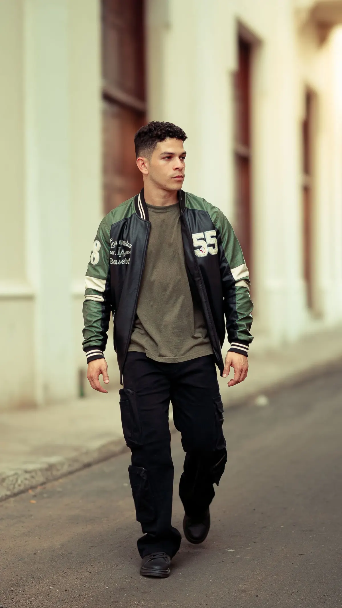 Pria berjalan di jalan kota memakai jaket varsity hitam berlengan hijau tua, kaus hijau zaitun, celana kargo hitam bersaku samping, dan sepatu hitam.