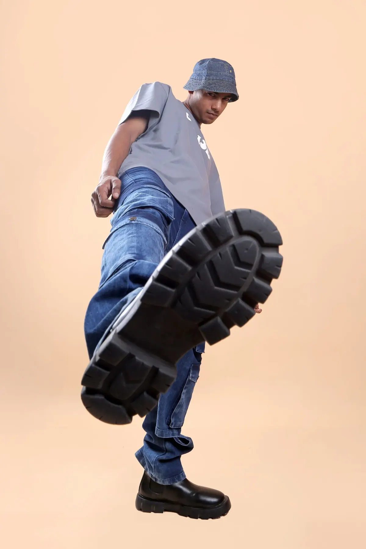 Foto studio dari sudut rendah: pria memakai topi bucket denim, kaus abu-abu longgar bergambar, celana denim biru bersaku kargo, dan boots hitam bersol tebal yang diangkat ke arah kamera.