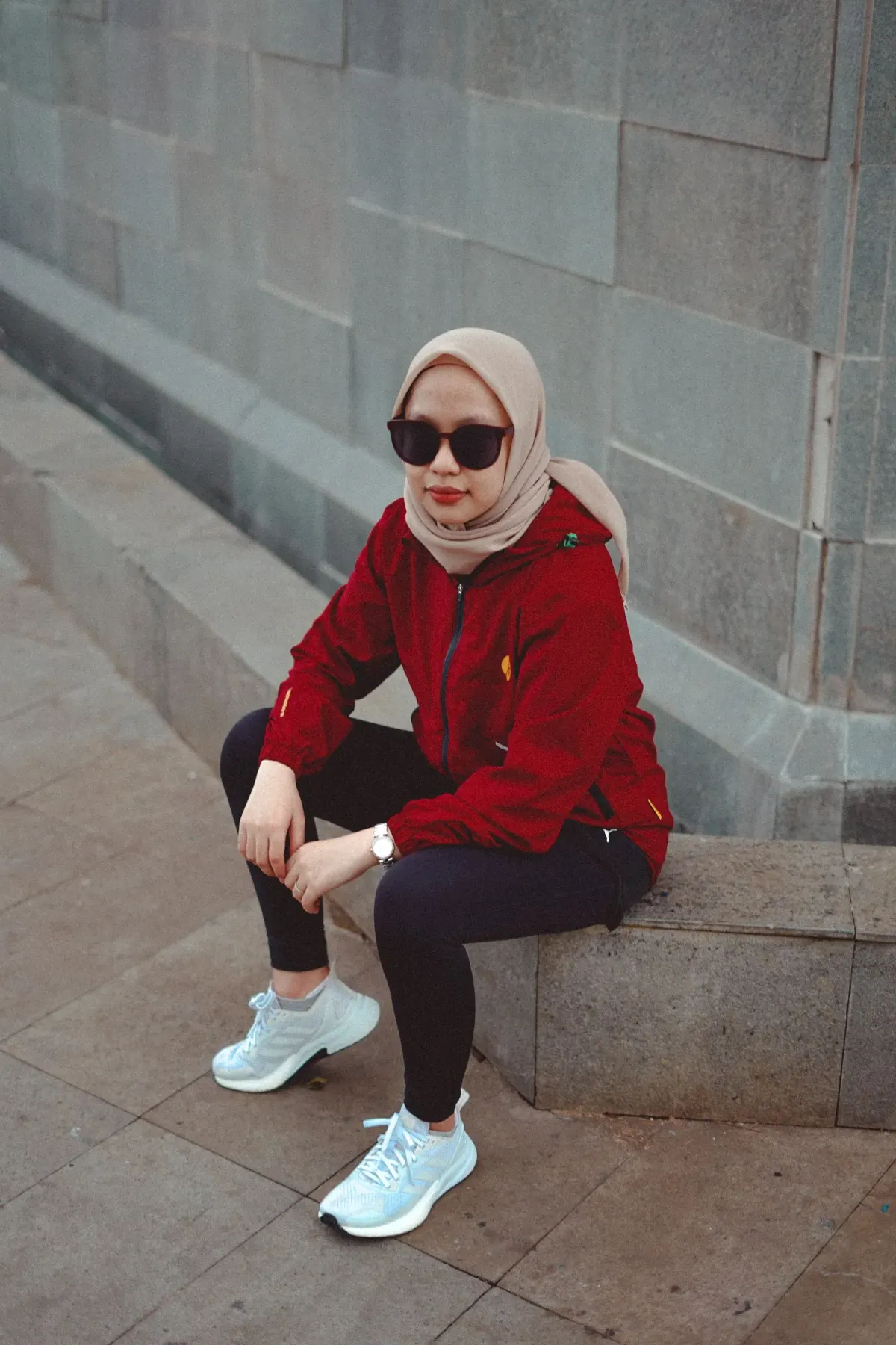 Perempuan berhijab krem dan berkacamata hitam duduk di bangku beton, memakai jaket merah berritsleting, legging hitam, dan sneakers bergaya lari berwarna biru muda pucat dengan sol tebal putih.