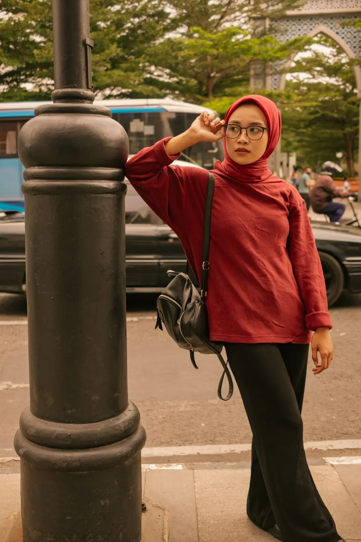 Perempuan berhijab terakota dan berkacamata memakai sweter merah longgar, celana hitam lebar, dan ransel hitam, bersandar di tiang lampu jalan.