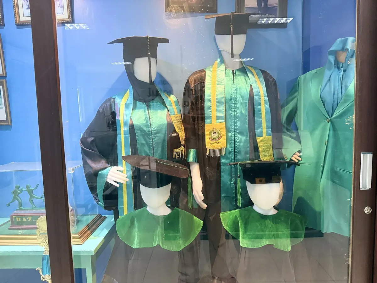 Etalase kaca berisi manekin bertoga hitam dengan topi toga, selempang hijau bergaris kuning yang menutup bagian depan badan, dan kerah hijau, di sebuah museum.