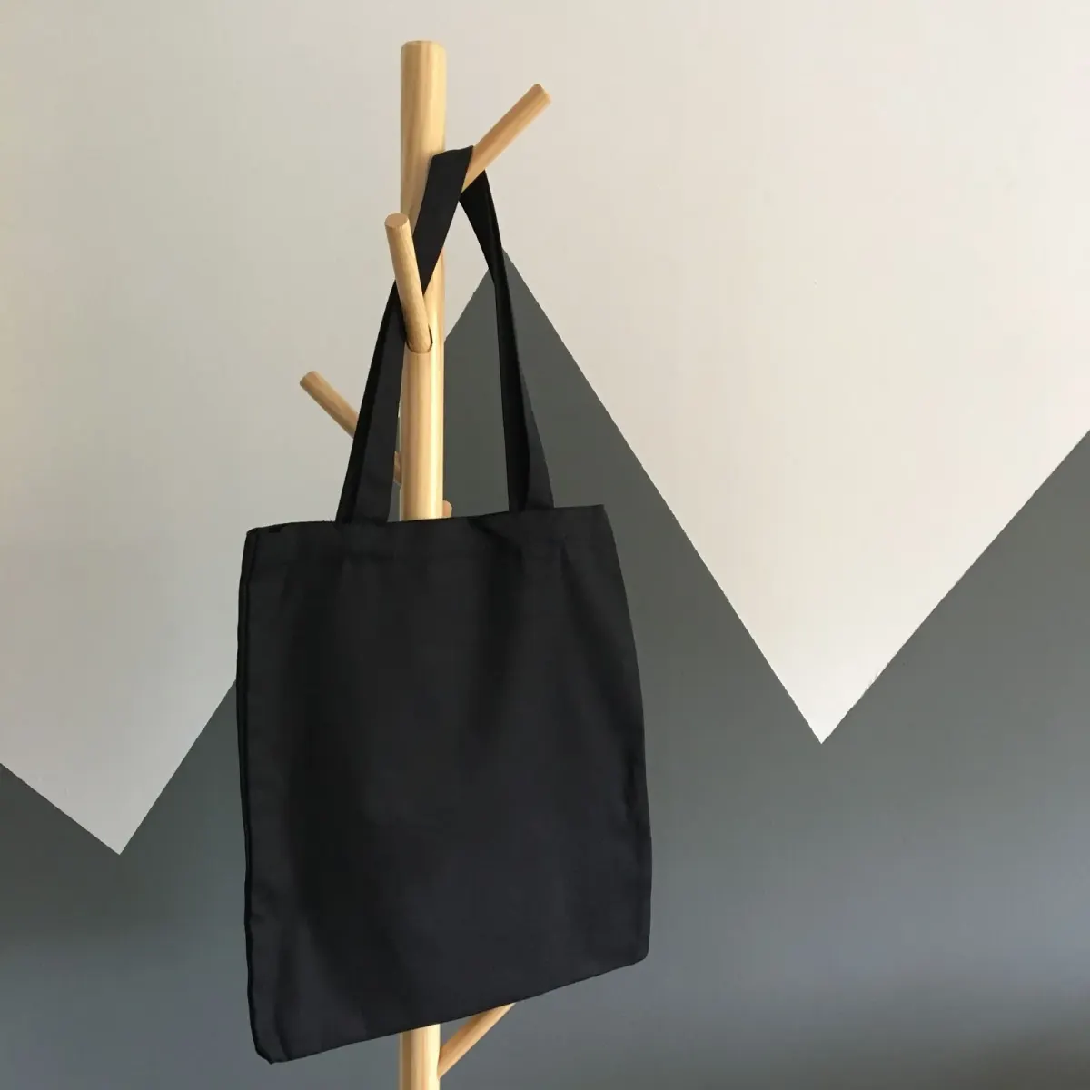 Tote bag kain hitam polos tergantung di tiang gantungan kayu, latar dinding putih dan abu-abu bermotif segitiga.