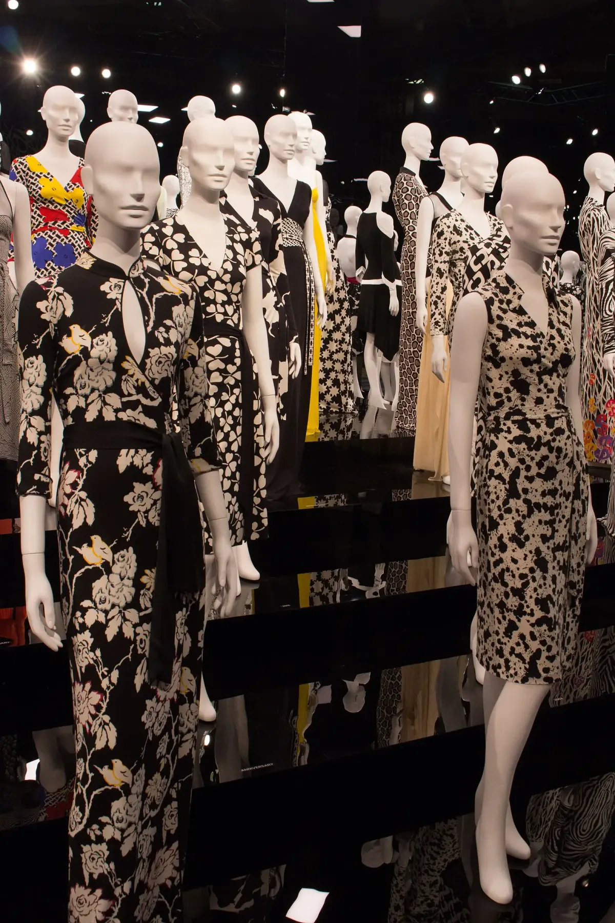 Deretan manekin putih memakai wrap dress bermotif bunga, macan tutul, dan geometris dalam pameran berlatar gelap.